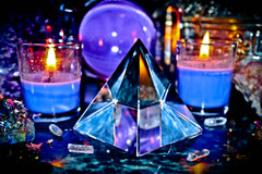 A charging crystal pyramid with a reflective surface, surrounded by candles and other metaphysical items on a dark background.